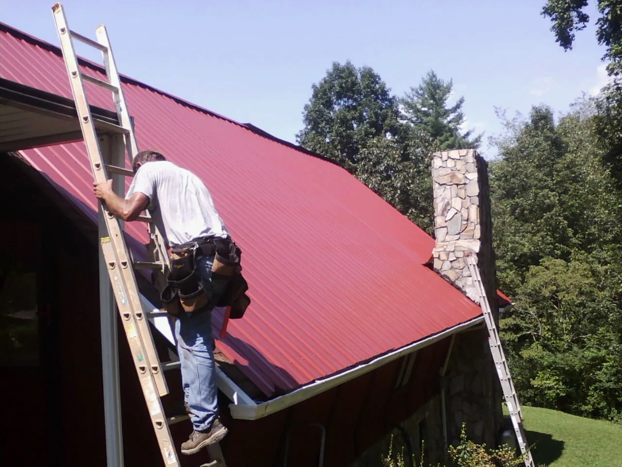 Roofer on a ladder beside a red metal roof and stone chimney — Soffit & Fascia Repair by Roofing Brothers in Curwensville